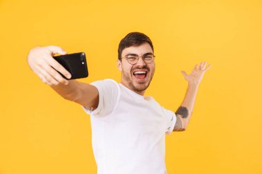 Gözlüklü neşeli genç adamın cep telefonuyla selfie çektiği ve sarı arka planda izole bir şekilde gülümsediği fotoğraf.