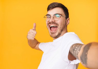 Gözlüklü neşeli bir gencin selfie çekerken ve sarı arkaplanda baş parmağını gösterirken çekilmiş bir fotoğraf.