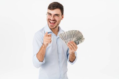 Gözlüklü, elinde dolar olan ve beyaz arka planda izole edilmiş kamerayı işaret eden mutlu bir adamın fotoğrafı.