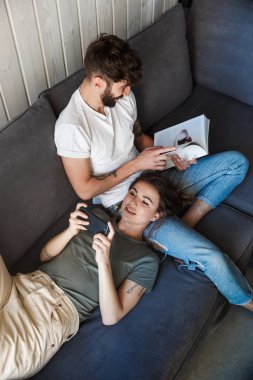Oturma odasındaki kanepede dinlenen gülümseyen genç çift kitap okuyan adam, cep telefonu kullanan kadın.