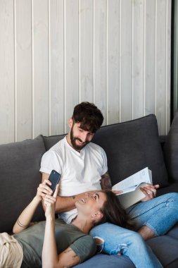 Oturma odasındaki kanepede dinlenen gülümseyen genç çift kitap okuyan adam, cep telefonu kullanan kadın.