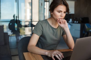 Evde masada otururken dizüstü bilgisayarda çalışan çekici, düşünceli genç bir kadın.