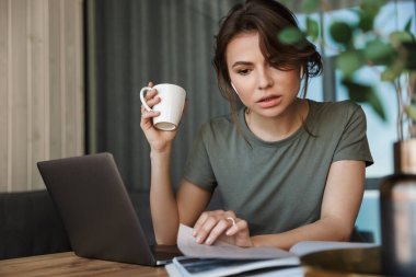 Evde oturmuş kahve içerken dizüstü bilgisayarda çalışan çekici, düşünceli genç bir kadın.