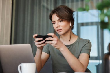 Evde dizüstü bilgisayarda çalışan, cep telefonu kullanan, kablosuz kulaklık takan çekici, düşünceli genç bir kadın.