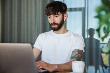 Mutfak masasında oturmuş, evde kahve içerken dizüstü bilgisayarda çalışan yakışıklı genç sakallı adam.