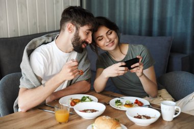 Güzel, mutlu, genç bir çift mutfakta sağlıklı bir kahvaltı yapıyor, cep telefonu kullanıyor.