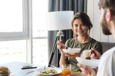 Güzel, mutlu, genç bir çift mutfakta sağlıklı bir kahvaltı yapıyor, sohbet ediyorlar.