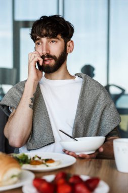Evde sağlıklı bir akşam yemeği yerken cep telefonu kullanan çekici genç sakallı adam.