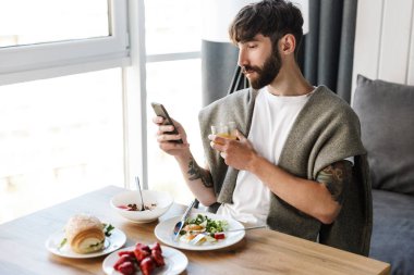 Evde sağlıklı bir akşam yemeği yerken cep telefonu kullanan çekici genç sakallı adam.