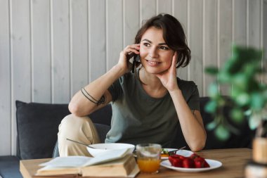 Çekici, gülümseyen genç bir kadın oturma odasında oturmuş cep telefonuyla konuşurken sağlıklı bir kahvaltı yapıyor.