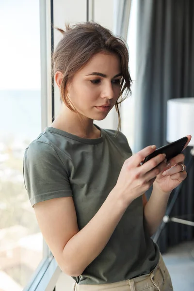 Attractive Young Woman Using Mobile Phone While Standing Window Indoors — Stock Photo, Image