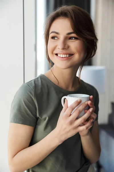 窓の外に立ちながらコーヒーカップを持つ魅力的な笑顔の若い女性 — ストック写真