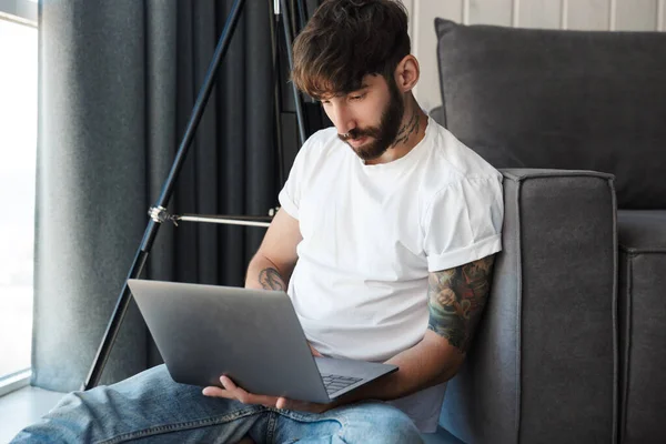 リビングルームの床にソファに座っている間 ラップトップコンピュータを使用してハンサムな自信のある若いひげそりの男 — ストック写真