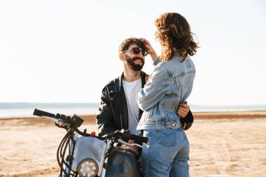 Çekici genç mutlu çift güneşli plajda motosiklete yaslanırken kucaklaşıyorlar.