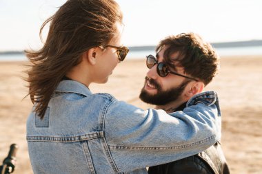 Çekici genç mutlu çift güneşli plajda motosiklete yaslanırken kucaklaşıyorlar.