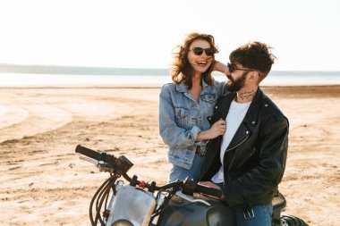 Çekici genç mutlu çift güneşli plajda motosiklete yaslanırken kucaklaşıyorlar.