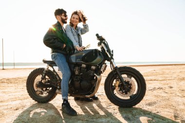 Çekici genç mutlu çift güneşli plajda motosiklete yaslanırken kucaklaşıyorlar.