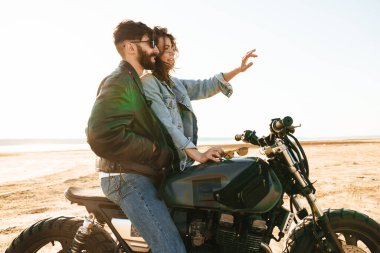 Çekici genç mutlu çift güneşli plajda motosiklete yaslanırken kucaklaşıyorlar.