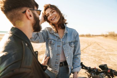 Çekici genç mutlu çift güneşli plajda motosiklete yaslanırken kucaklaşıyorlar.