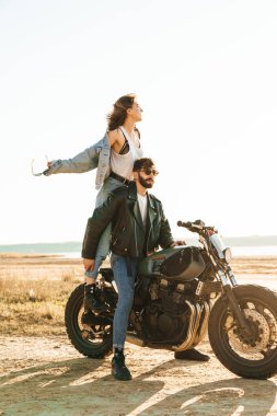 Çekici genç mutlu çift güneşli plajda motosiklete yaslanırken kucaklaşıyorlar.
