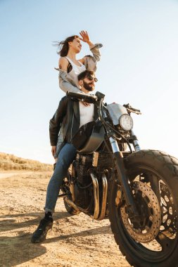 Çekici genç mutlu çift güneşli plajda motosiklete yaslanırken kucaklaşıyorlar.