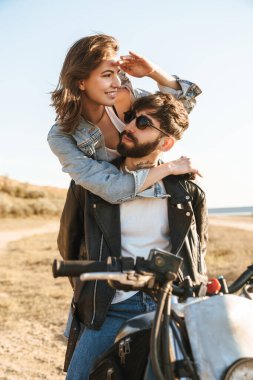 Çekici genç mutlu çift güneşli plajda motosiklete yaslanırken kucaklaşıyorlar.