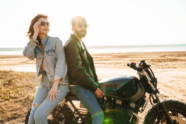 Çekici genç mutlu çift güneşli plajda motosiklete yaslanırken kucaklaşıyorlar.