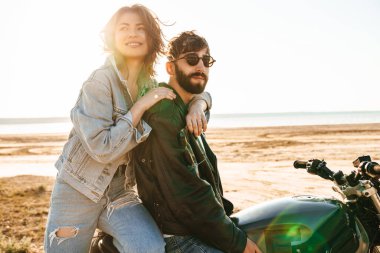 Güneşli sahilde motosiklet üzerinde oturan ceketli güzel Yong çifti sarılıyorlar.