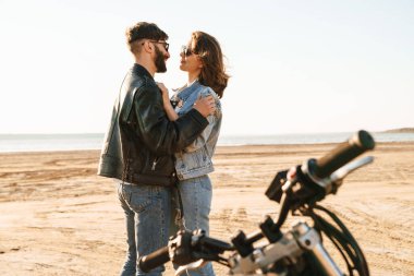 Ceket giyen genç ve güzel Cople güneşli sahilde motosikletin yanında duruyor.