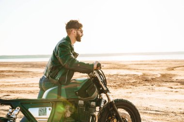 Deri ceket giyen yakışıklı genç adam güneşli sahilde motosikletle yürüyor.