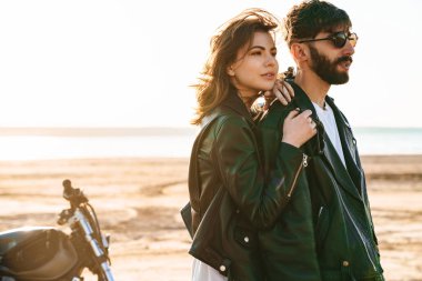 Çekici genç çift güneşli sahilde motosikletin yanında dururken kucaklaşıyorlar.