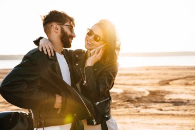 Çekici genç çift güneşli sahilde motosikletin yanında dururken kucaklaşıyorlar.
