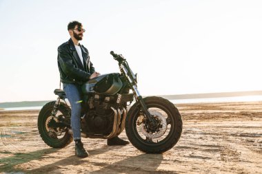 Kumsalda motosiklette oturan deri ceket giyen yakışıklı genç sakallı adam.