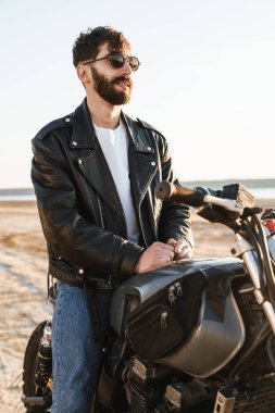 Kumsalda motosiklette oturan deri ceket giyen yakışıklı genç sakallı adam.