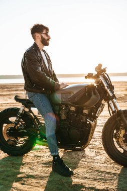 Kumsalda motosiklette oturan deri ceket giyen yakışıklı genç sakallı adam.