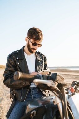 Deri ceketli yakışıklı genç adam plajda motosiklette oturuyor, cep telefonu kullanıyor.