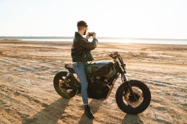 Deri ceketli yakışıklı genç adam güneşli sahilde motosiklette oturmuş manzaranın fotoğrafını çekiyor.