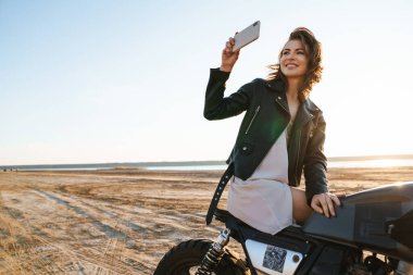 Deri ceketli güzel bir genç kız güneşli sahilde motosiklette oturmuş, selfie çekiyor.