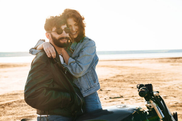 Attractive young happy couple embracing while leaning on a motorbike at the sunny beach