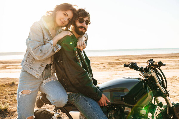Beatiful yong couple wearing jackets sitting on a motorbike at the sunny beach, hugging