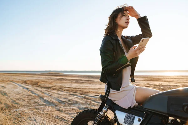携帯電話を使用して 晴れたビーチでバイクに座って革のジャケットを身に着けている美しい若い女の子 — ストック写真