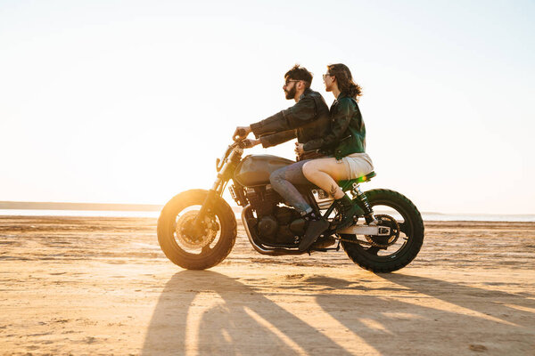 Side view of a beautiful young stylish couple enjoying ride on a motorbike at the beach