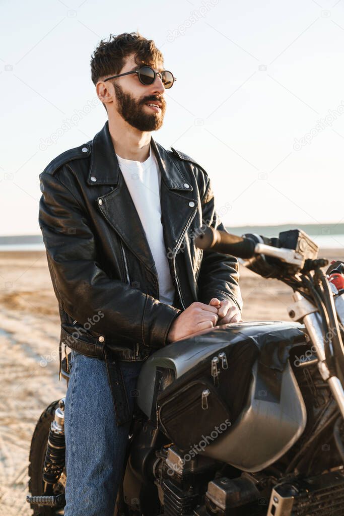 Handsome young bearded man wearing leather jacket sitting on a motorbike at the beach
