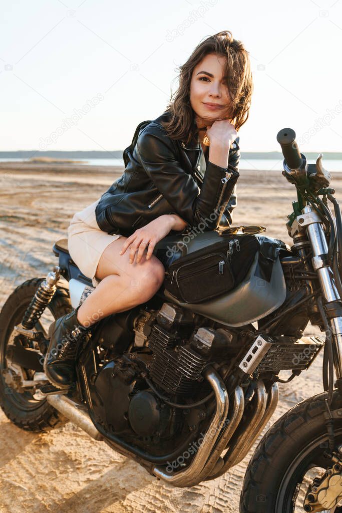Beautiful young woman wearing lether jacket sitting on a motorbike at the beach