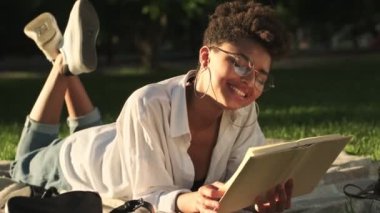 Gülümseyen Afro-Amerikan bir kadın parkta piknik sırasında kitap okuyor.