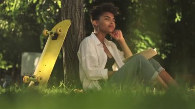 Güzel, Afro-Amerikan bir kadın elinde bir kitapla ağacın yanındaki parkta otururken düşünüyor.