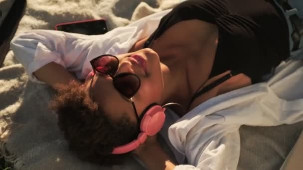 一位面带微笑的年轻非洲裔美国女人的头像是在公园里躺下野餐时 用耳机听音乐 — 图库视频影像