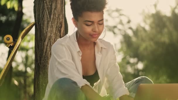 一位年轻美丽的非洲姑娘正坐在公园的草地上 用她的笔记本电脑 — 图库视频影像