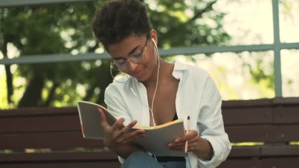 Positiv Ung Afroamerikansk Kvinne Med Øretelefoner Ser Notatboken Sin Parken – stockvideo