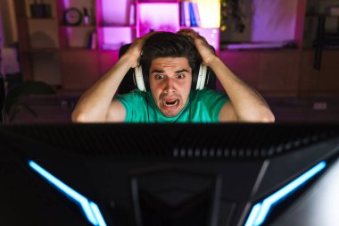 Kulaklıklı mutsuz bir gencin evde bilgisayar oyunu oynarken kafasını tutuşu.